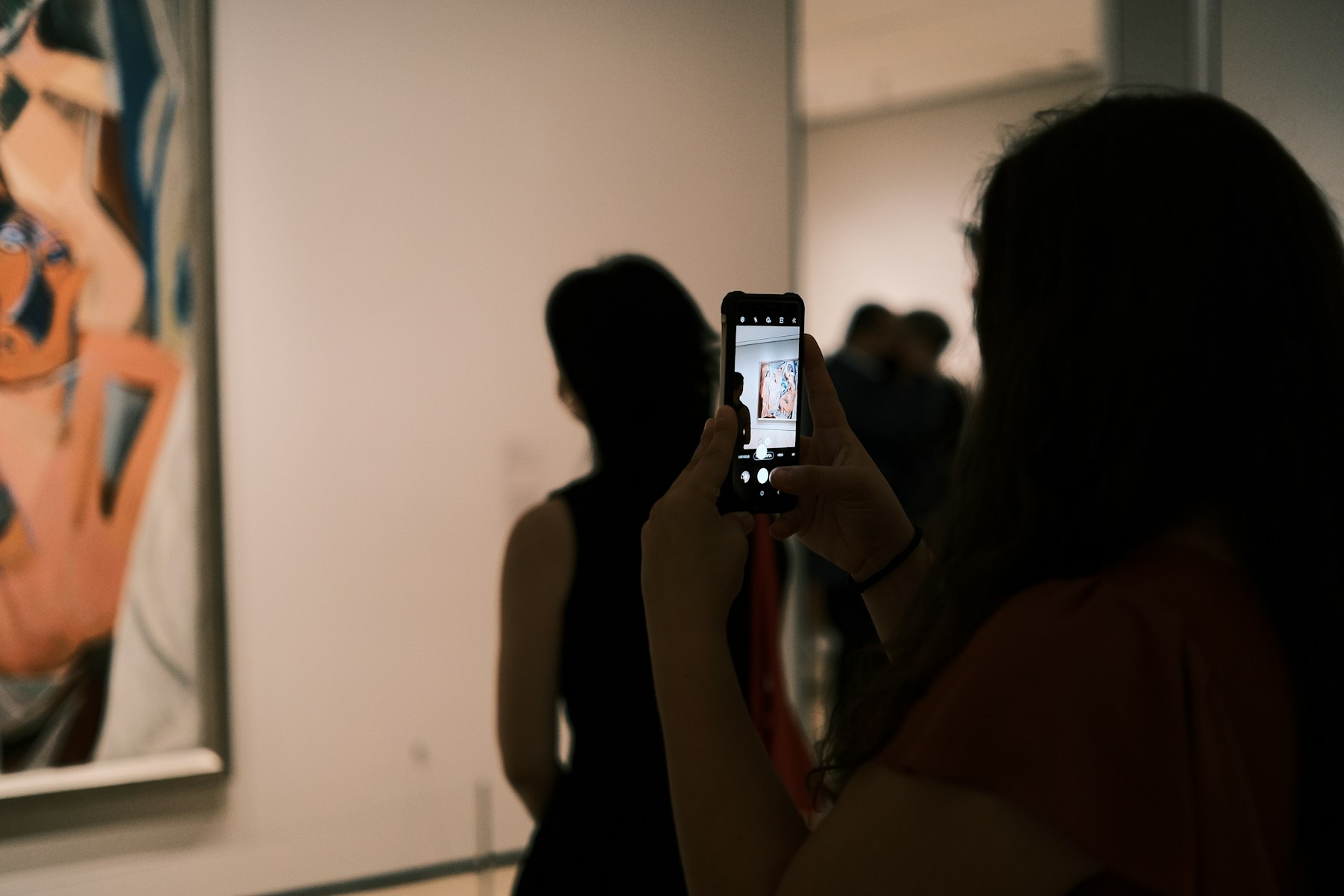 A visitor in a dim gallery photographs a painting with her phone, held at an angle, under spotlights.