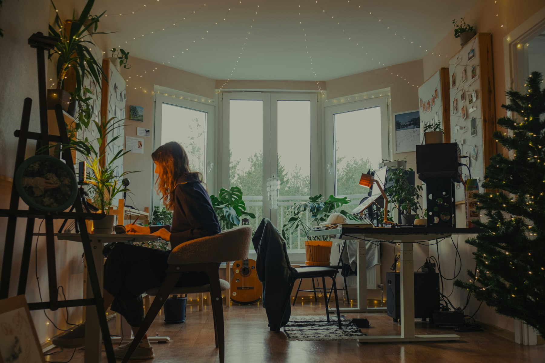 An artist working at a laptop in a warm studio with an easel, plants and drawings pinned to the walls.
