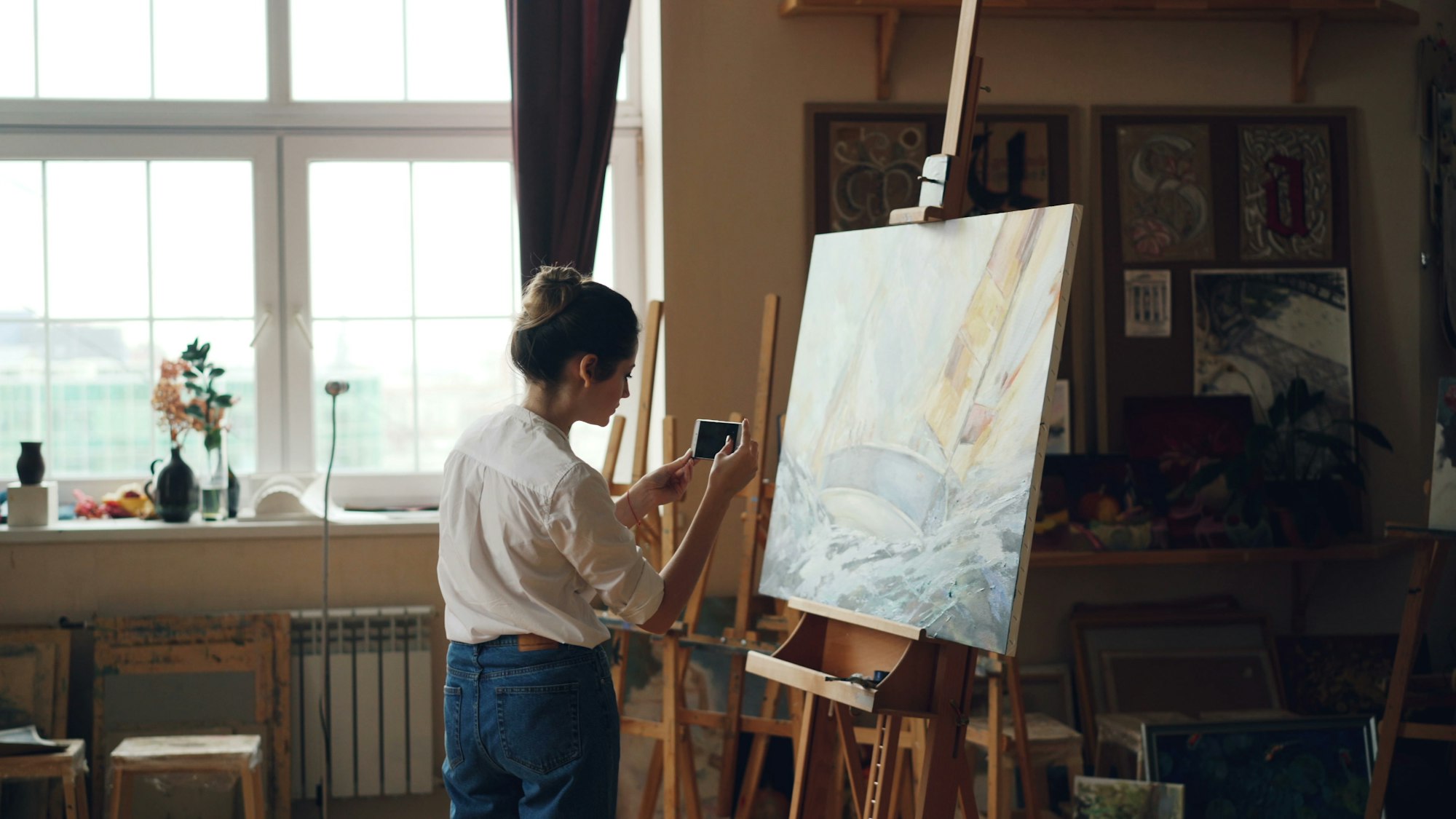 A painter in a sunlit studio photographs her canvas of a sailing boat with her phone.